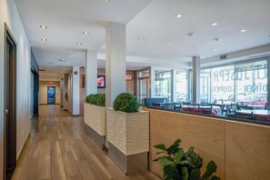 Un vestíbulo de hotel con mostrador de recepción, piso de madera, plantas en macetas y ventanas amplias.