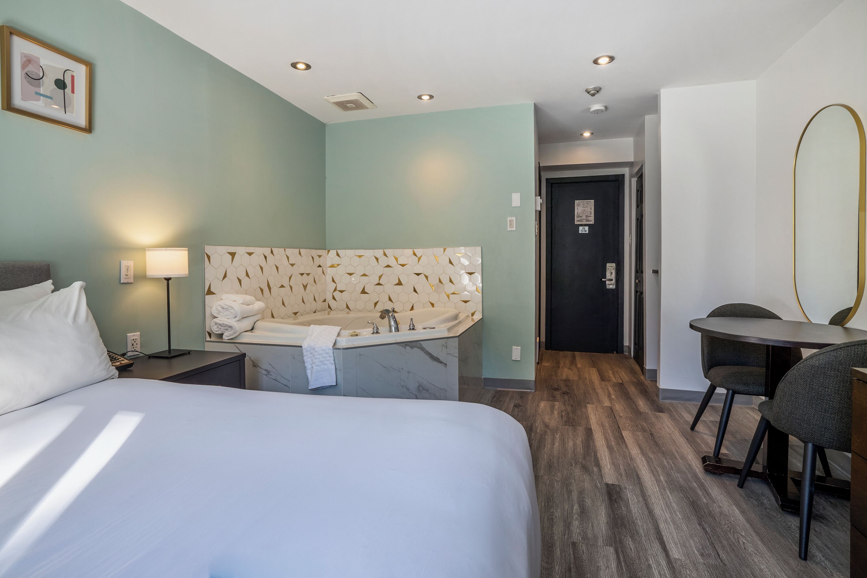 A hotel room with a bed, a desk with a chair, a small table, a wall-mounted mirror, a bathtub, and a door leading to another room.