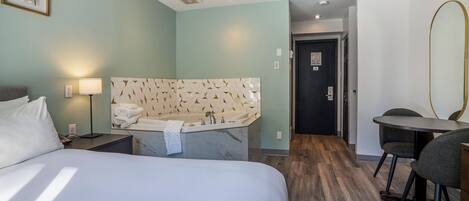A hotel room with a bed, a desk with a chair, a small table, a wall-mounted mirror, a bathtub, and a door leading to another room.