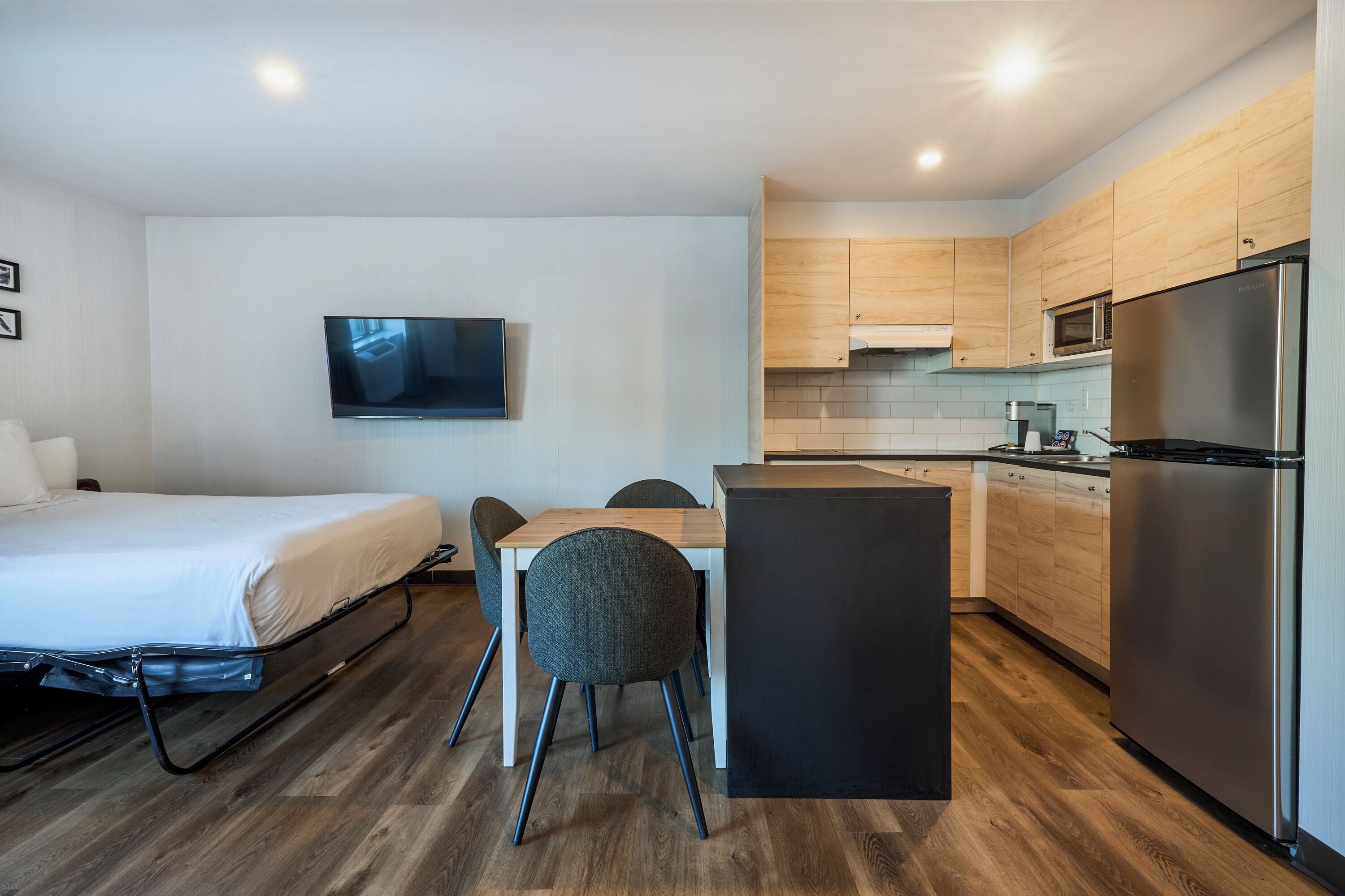 A compact hotel room with a bed, a dining table with chairs, a TV mounted on the wall, a kitchenette with a refrigerator, and wooden flooring.