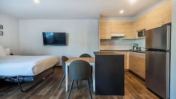Habitación de hotel compacta con cama, una mesa de comedor con sillas, televisor montado en la pared, kitchenette con refrigerador y piso de madera.