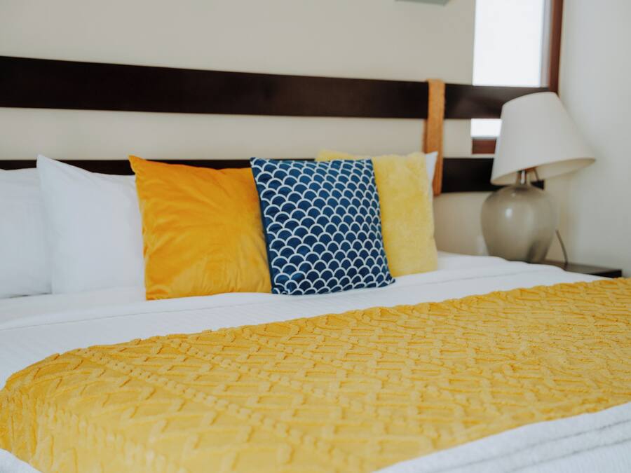 Una cama bien hecha con una colcha amarilla con textura, almohadas blancas y una almohada azul estampada.