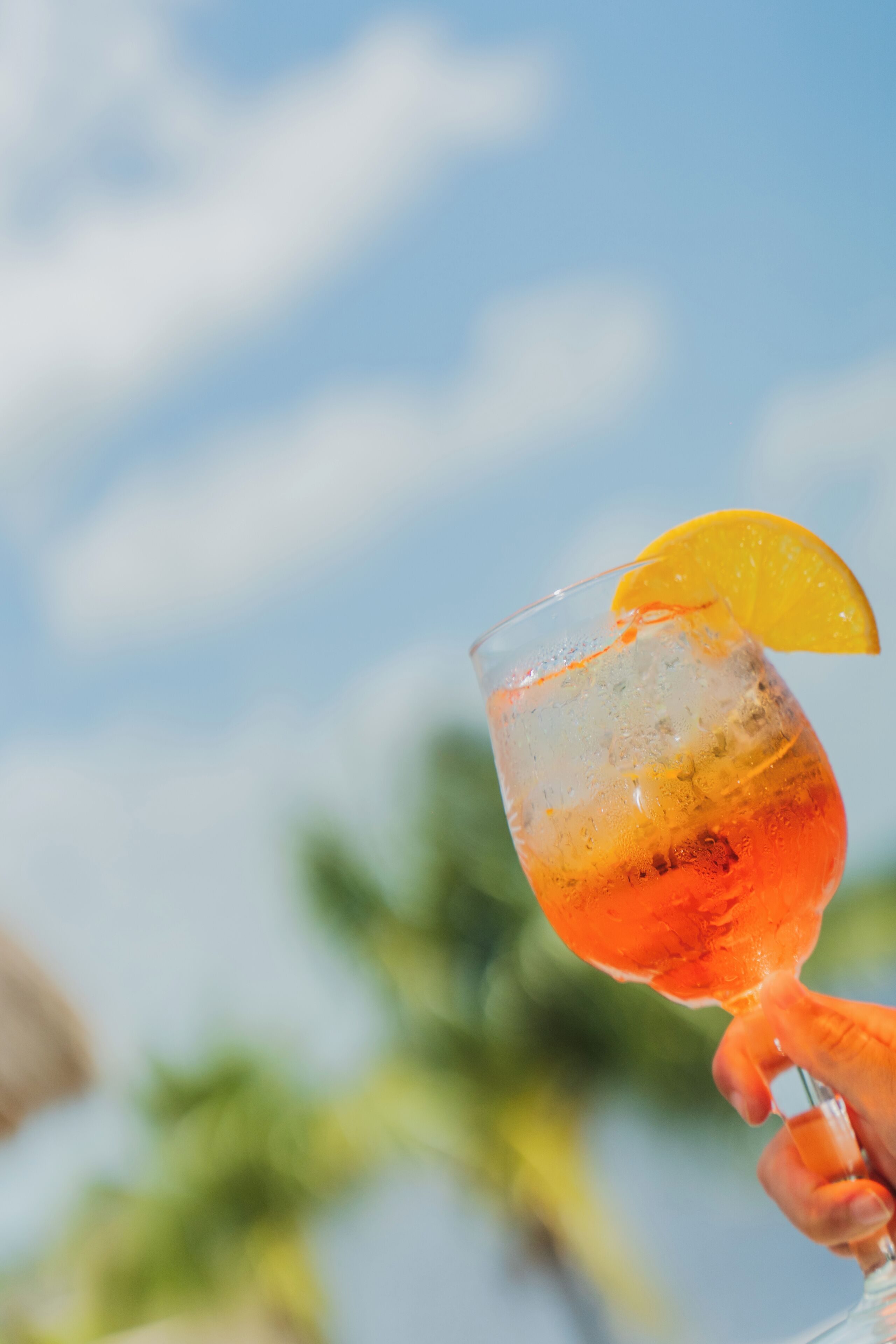 A hand holding a cocktail glass with a slice of orange garnish against a clear sky.