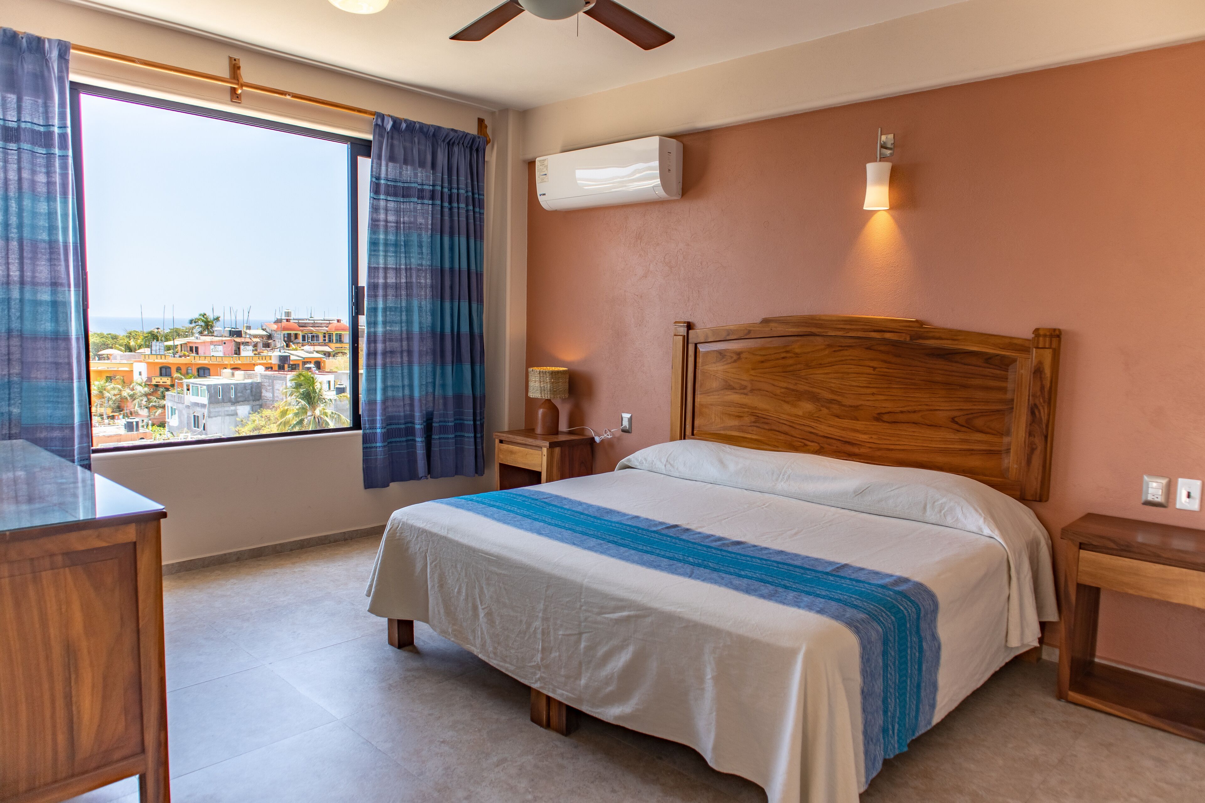 A bedroom with a large window, a wooden headboard, a bed with a white and blue striped bedspread, a nightstand, and an air conditioning unit on the wall.