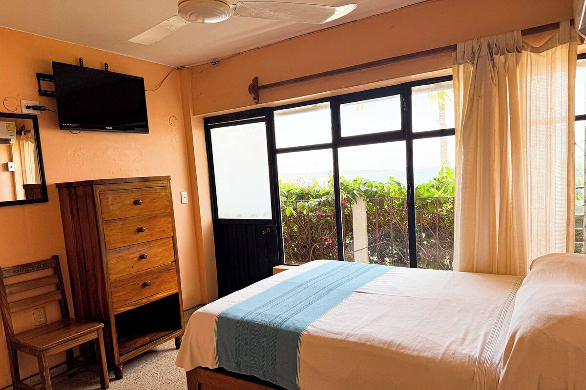 A bedroom with a bed, a wooden dresser, a television, and a large window with a view of greenery and a body of water.