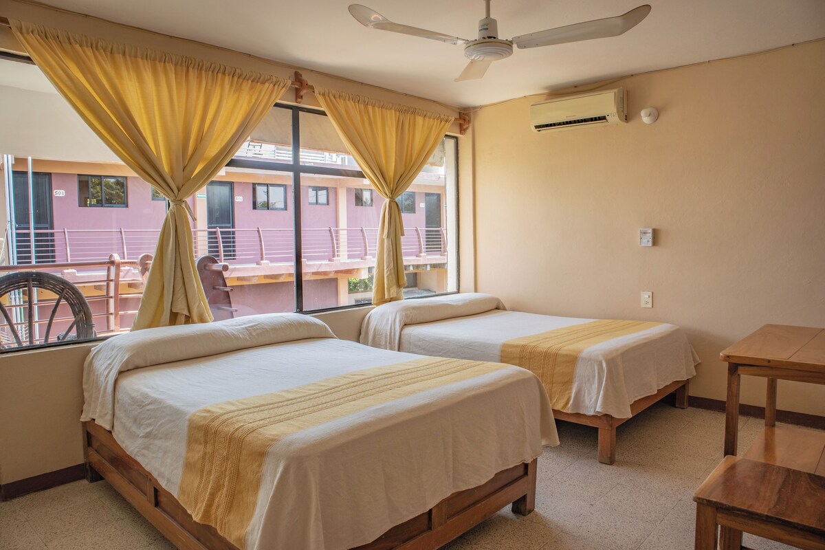 Habitación de hotel con dos camas individuales, ventilador de techo, aire acondicionado y vista a un edificio.