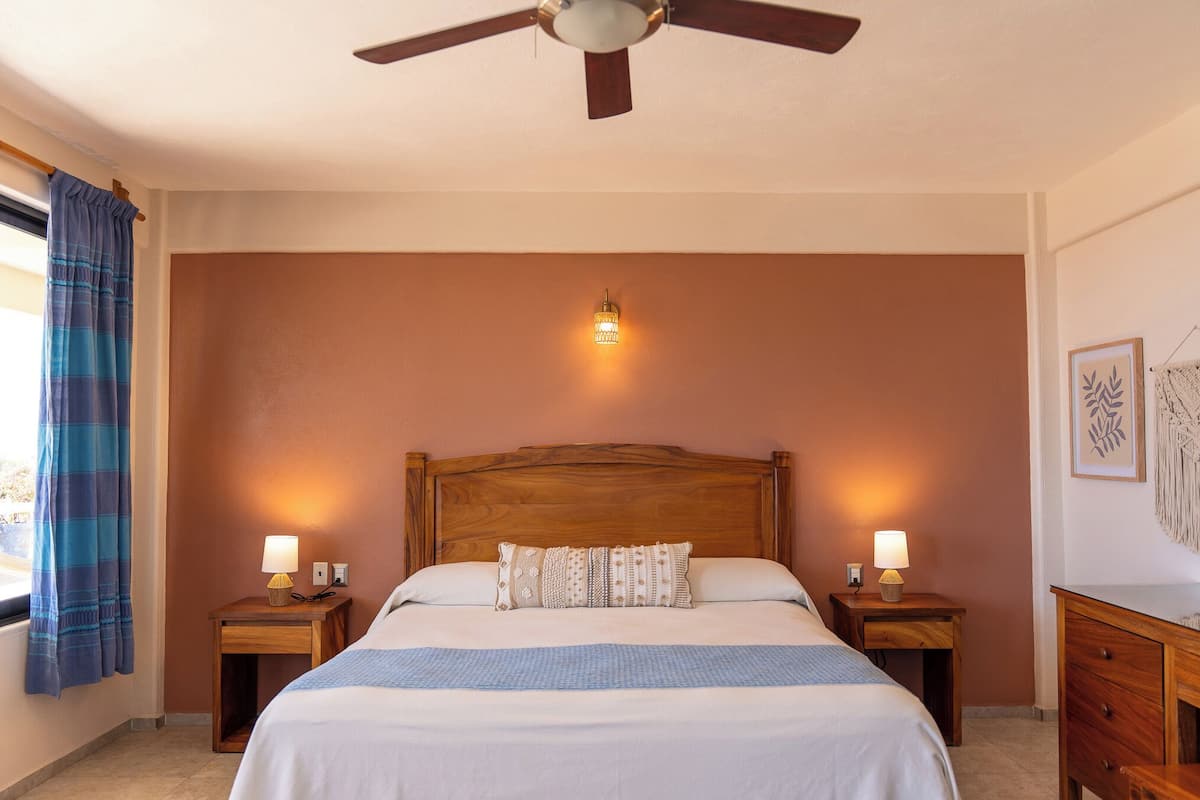 A hotel room with a wooden headboard, two bedside tables with lamps, a bed with a patterned pillow, a wall-mounted light, and a ceiling fan.