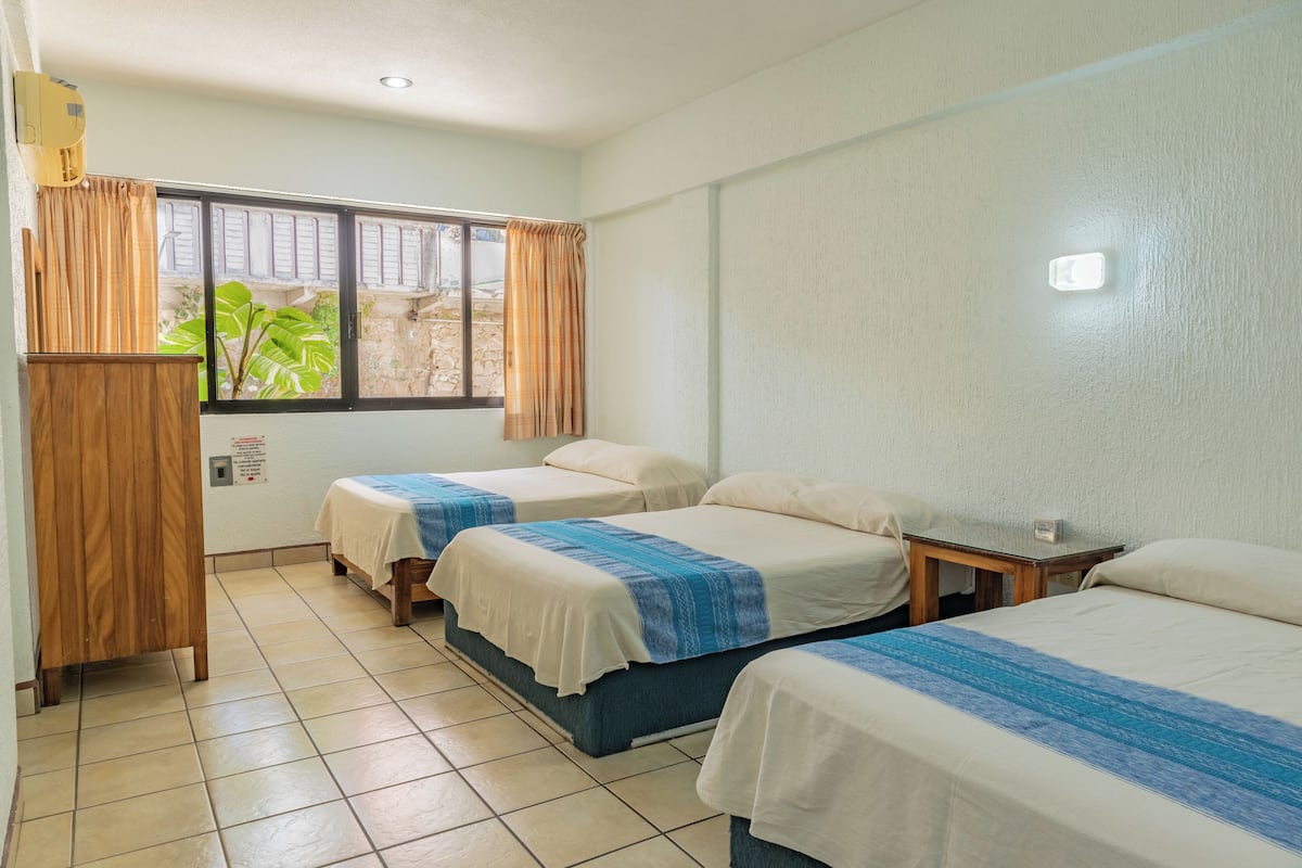 A hotel room with three single beds, a wooden wardrobe, a window with curtains, and a tiled floor.