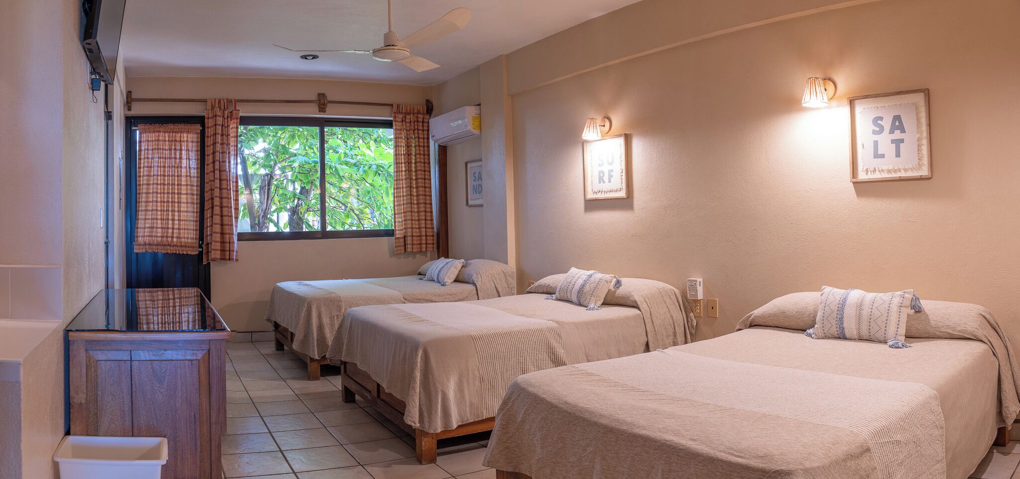 Habitación de hotel con tres camas, ventilador de techo, ventana con cortinas y una lámpara fijada a la pared.