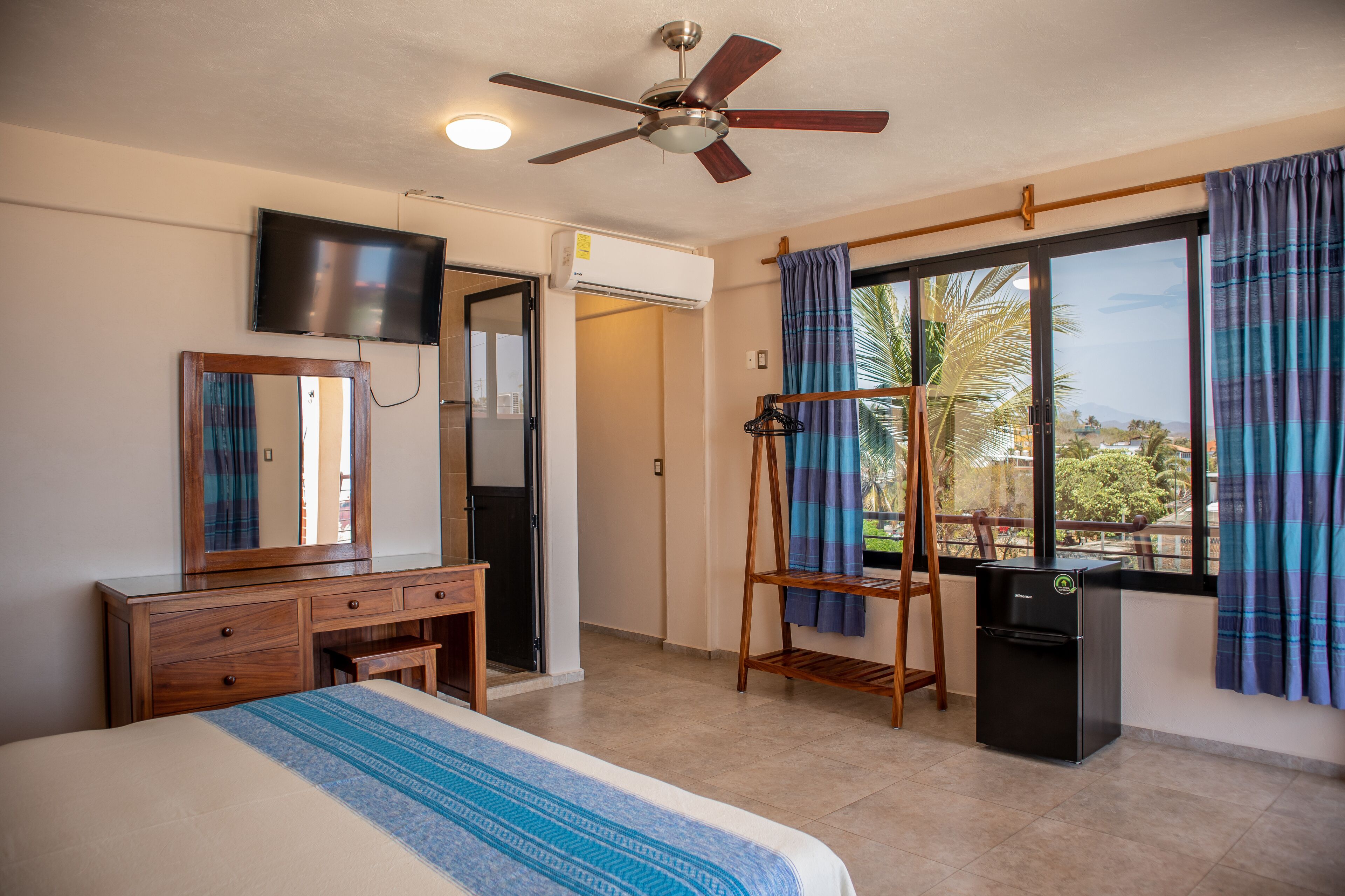 A hotel room with a bed, a dresser, a mirror, a television, a ceiling fan, a window with curtains, and a small refrigerator.