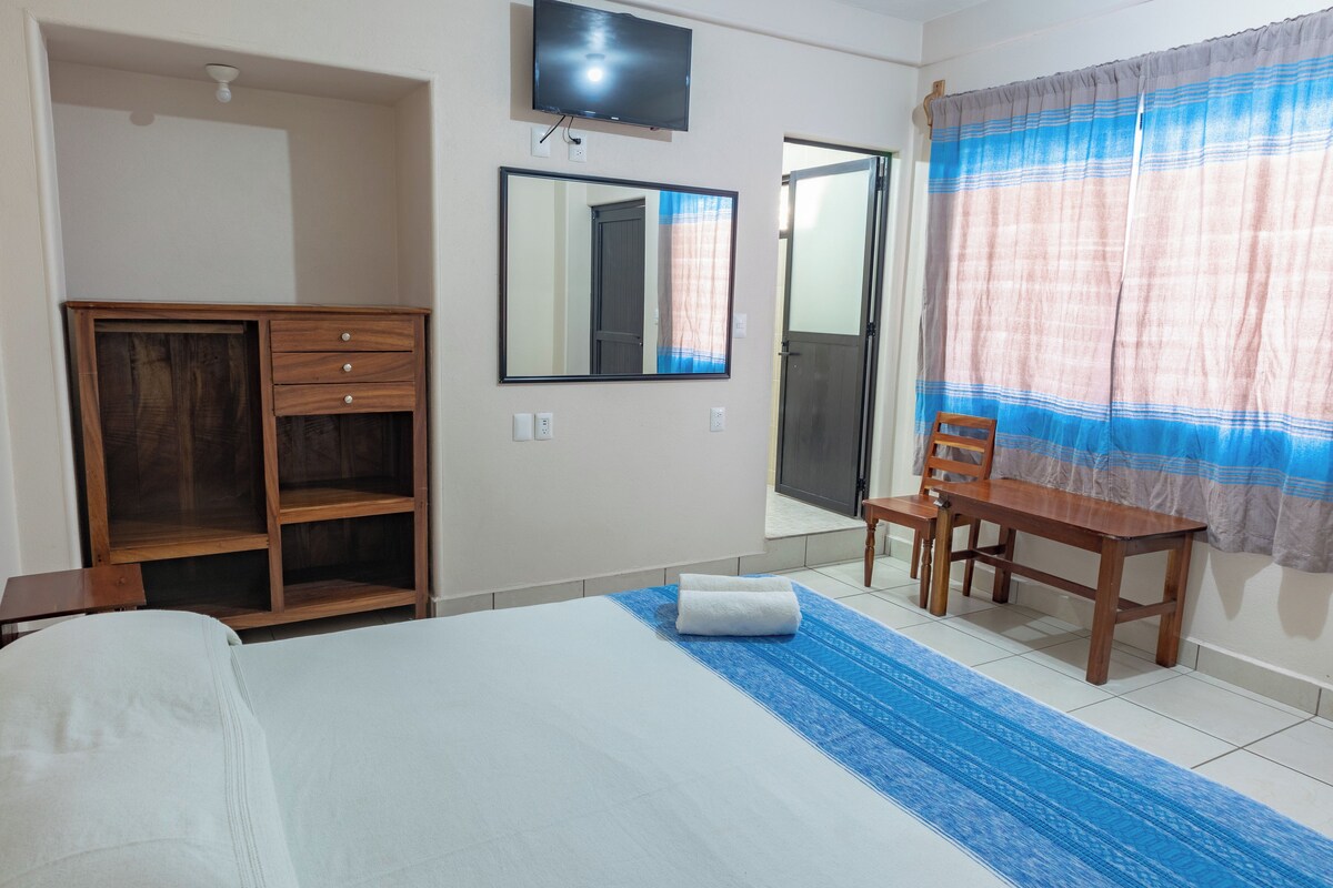 Habitación de hotel con cama, escritorio de madera, silla, televisor, armario y ventana con cortinas.