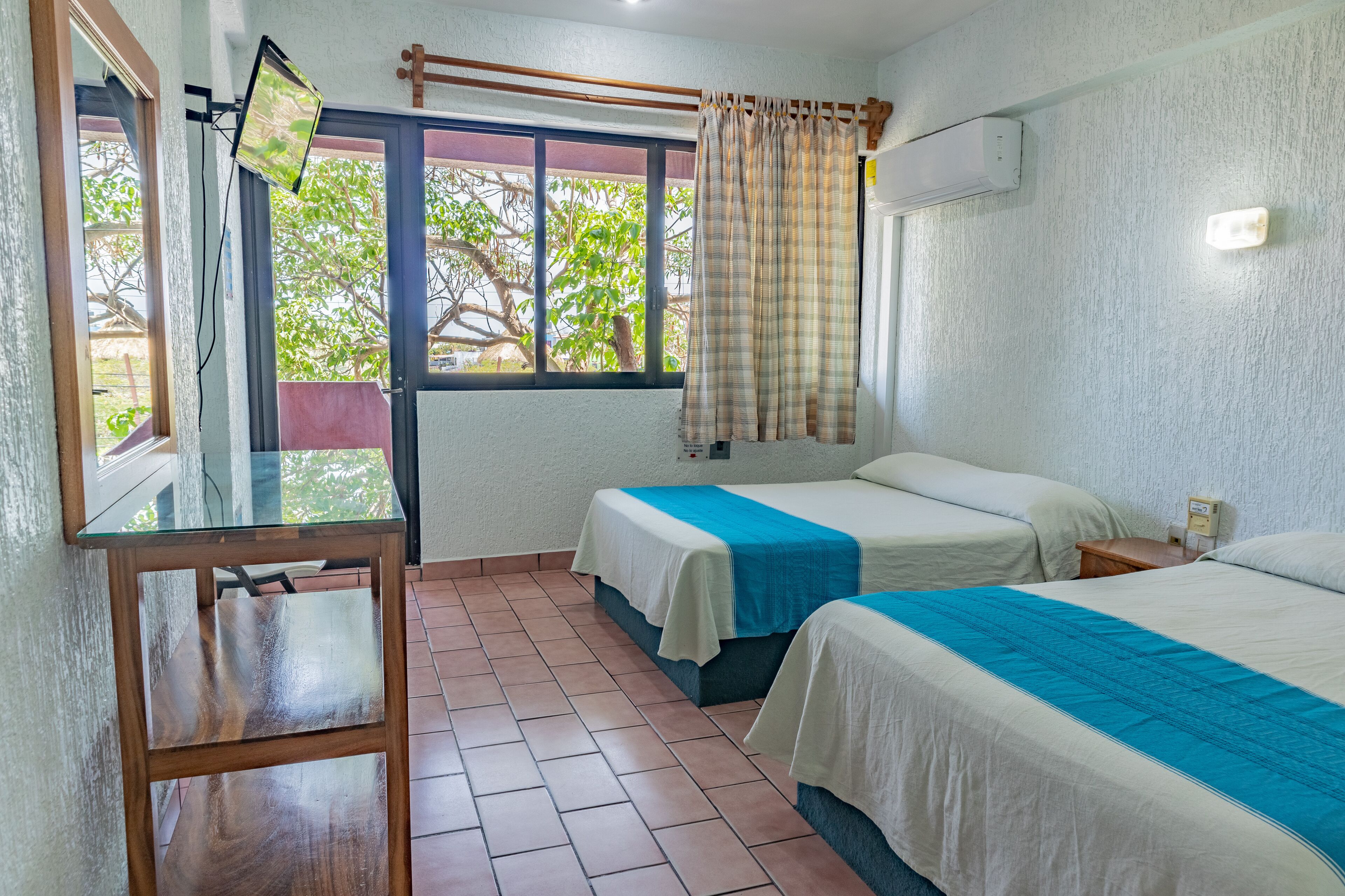 A room with two beds, a wooden table, a window with curtains, and an air conditioning unit.