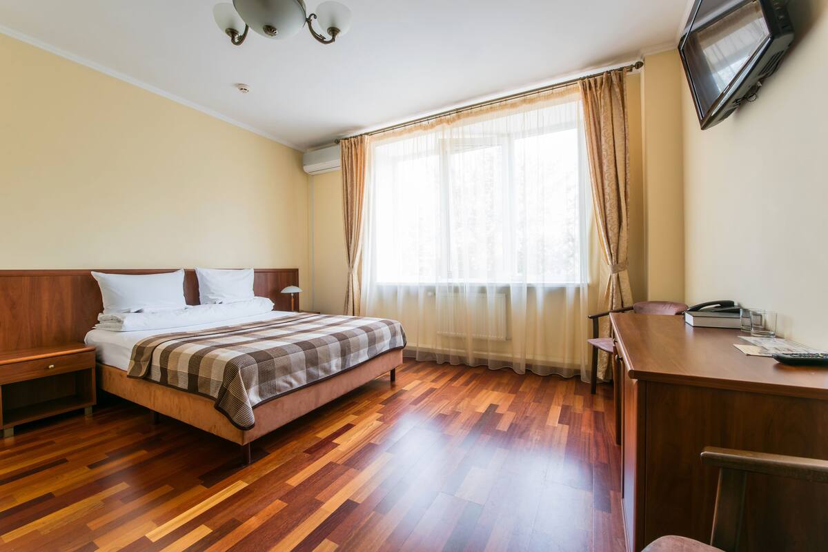 A hotel room with a wooden floor, a bed with a checkered blanket, a desk with a telephone, and a TV mounted on the wall.