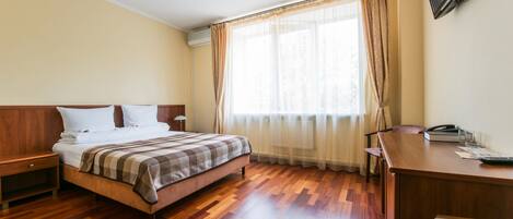A hotel room with a wooden floor, a bed with a checkered blanket, a desk with a telephone, and a TV mounted on the wall.