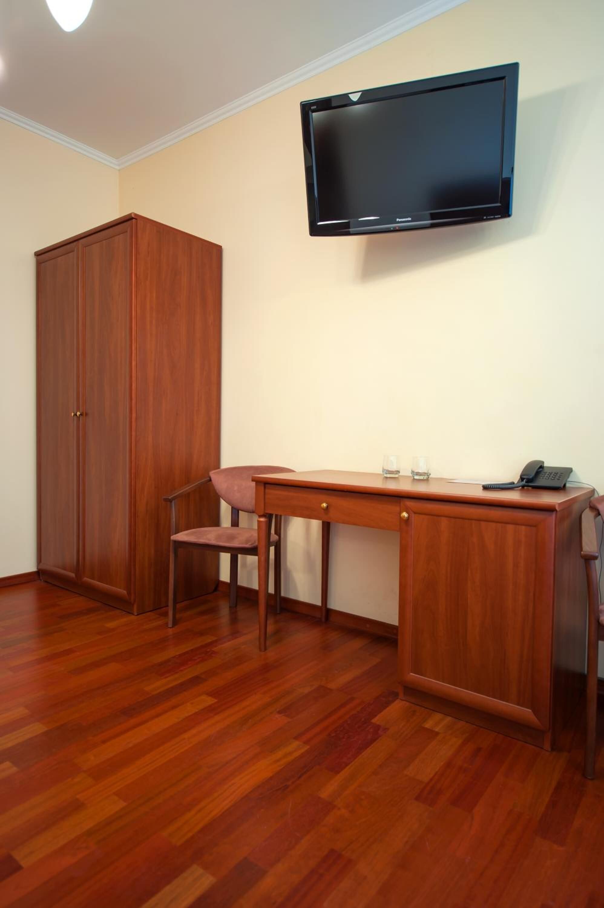 A room with a wooden desk, a chair, a wardrobe, and a mounted television.