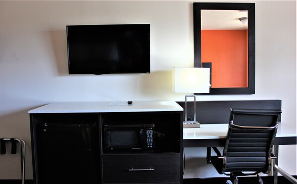 Habitación de hotel con televisión de pantalla plana montada en la pared, escritorio con silla, microondas y un mueble pequeño.