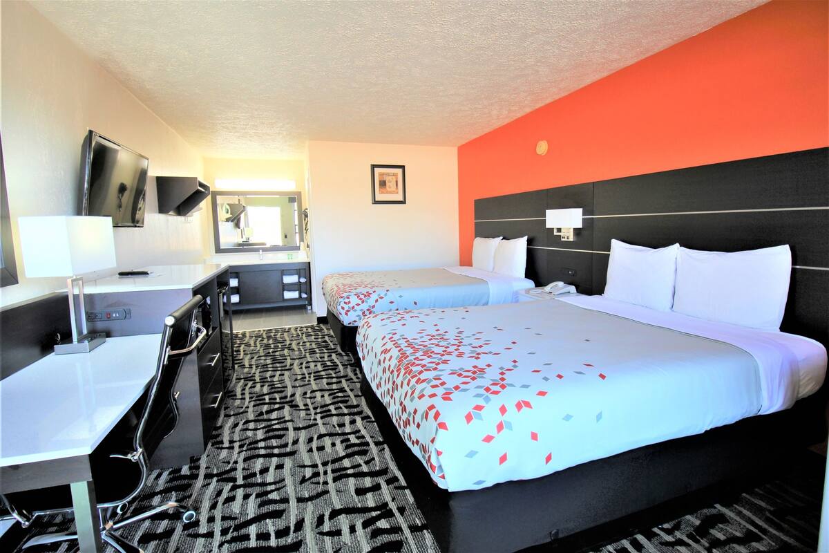 Quarto de hotel com duas camas, uma escrivaninha, uma cadeira, televisão e espelho no banheiro.