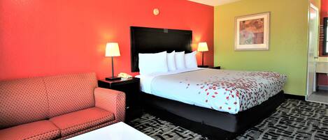 Una habitación de hotel con una cama, un sofá rojo, una mesa de centro blanca y una alfombra estampada.