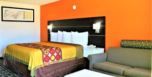 Quarto de hotel com parede laranja, cama com colcha estampada, cabeceira com iluminação, sofá verde e mesa de cabeceira com telefone.