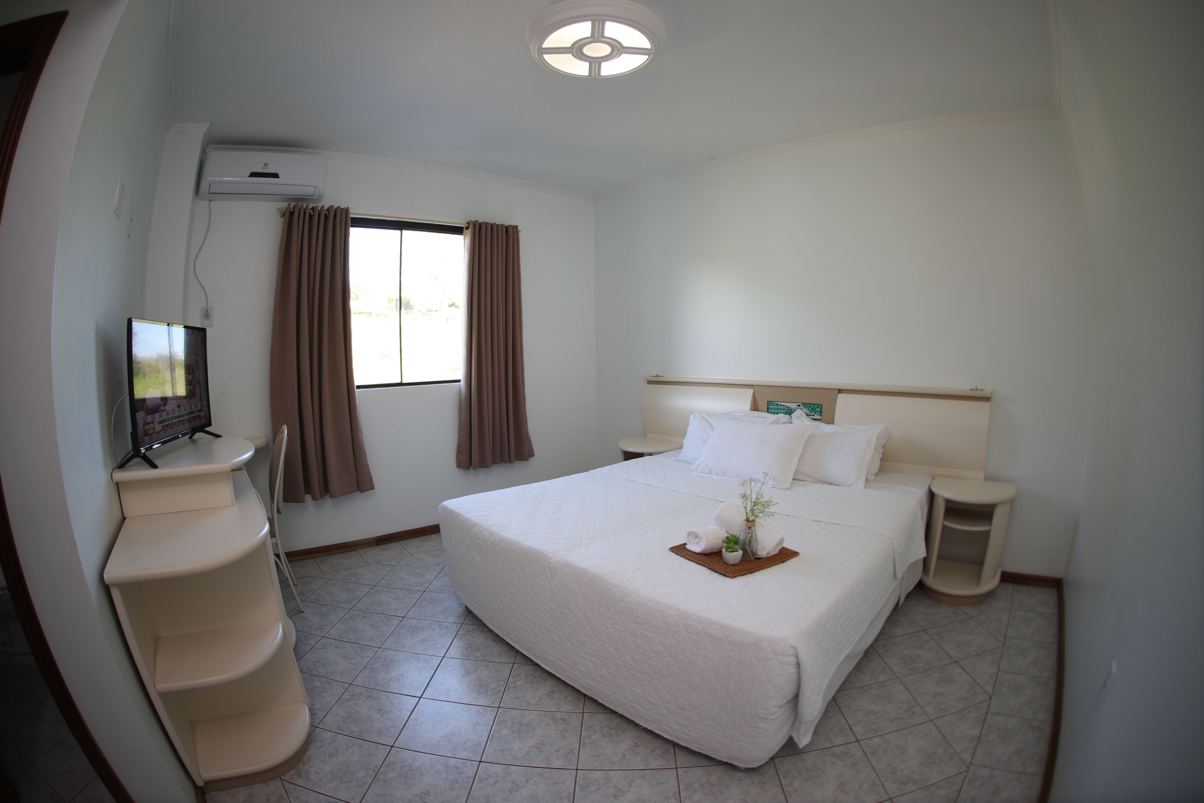 Quarto de hotel com cama, escrivaninha, cadeira, televisão e janela com cortinas.