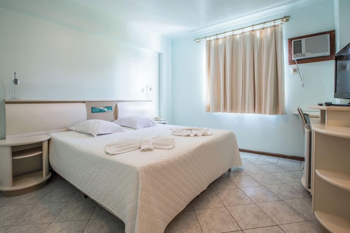 Una habitación de hotel con cama, mesitas de noche, televisor y ventana con cortinas.