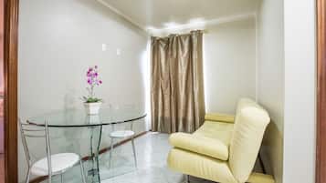 Um quarto pequeno e bem iluminado com uma mesa redonda de vidro, duas cadeiras, uma poltrona amarela, um vaso com planta e uma cortina.