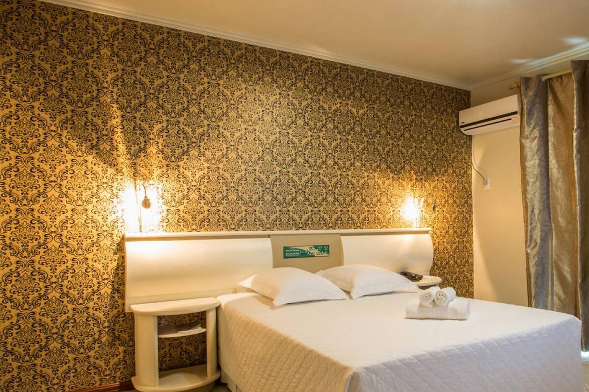 Habitación de hotel con cama, mesita de noche, aire acondicionado montado en la pared y una pequeña mesa con lámpara.