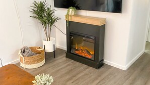 A living room with a flat-screen TV mounted on the wall, a fireplace with a visible fire, a wooden coffee table with magazines and a remote control, and a potted plant.