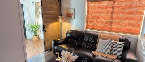 Sala de estar com sofá de couro preto, mesa de centro com objetos decorativos, tapete estampado e uma grande janela com persianas.