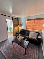 A living room with a black leather sofa, a coffee table with decorative items, a patterned rug, and a large window with blinds.