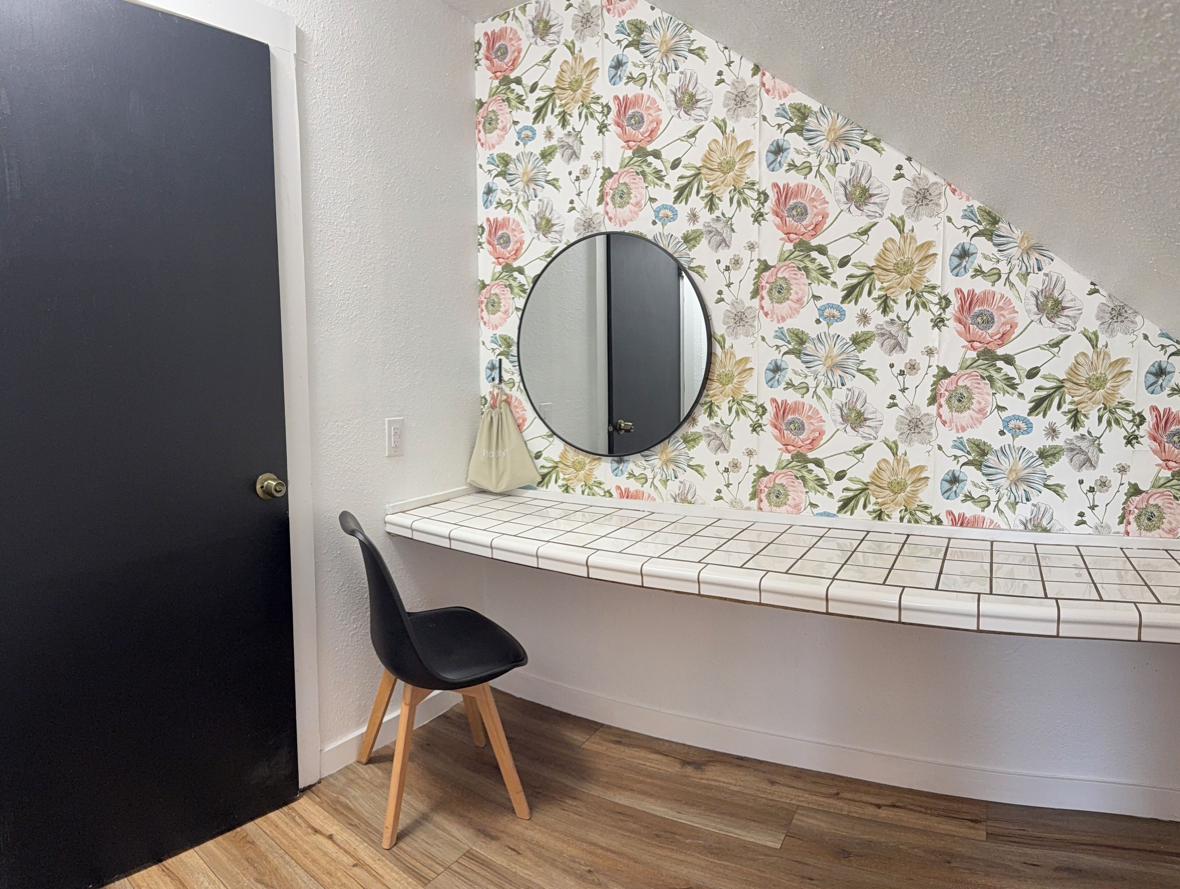 A corner bathroom with a white tiled vanity, a black door, a wooden chair, and a floral wallpaper.