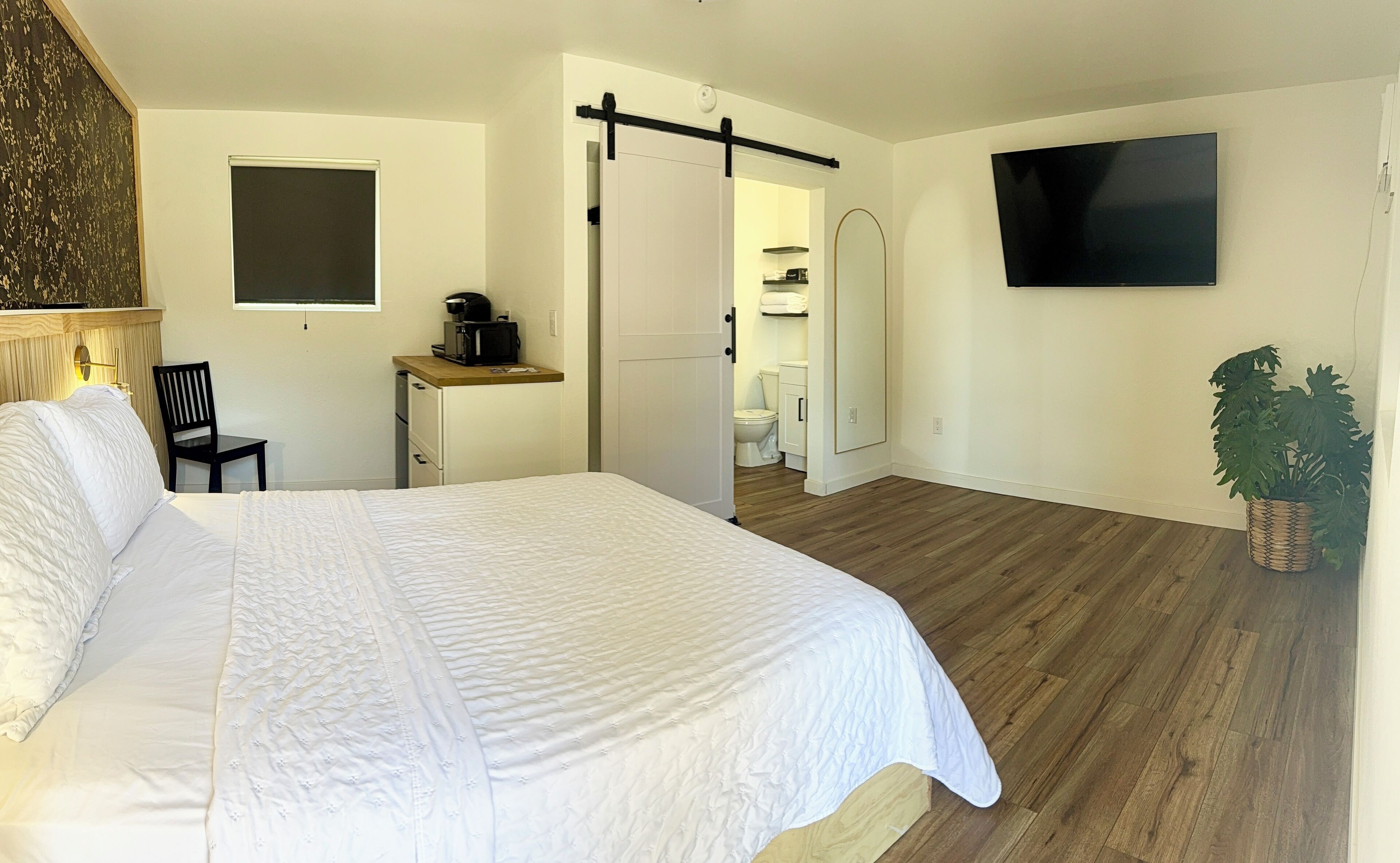 Habitación de hotel con una cama grande, un escritorio con cafetera, televisor montado en la pared y una planta en maceta.
