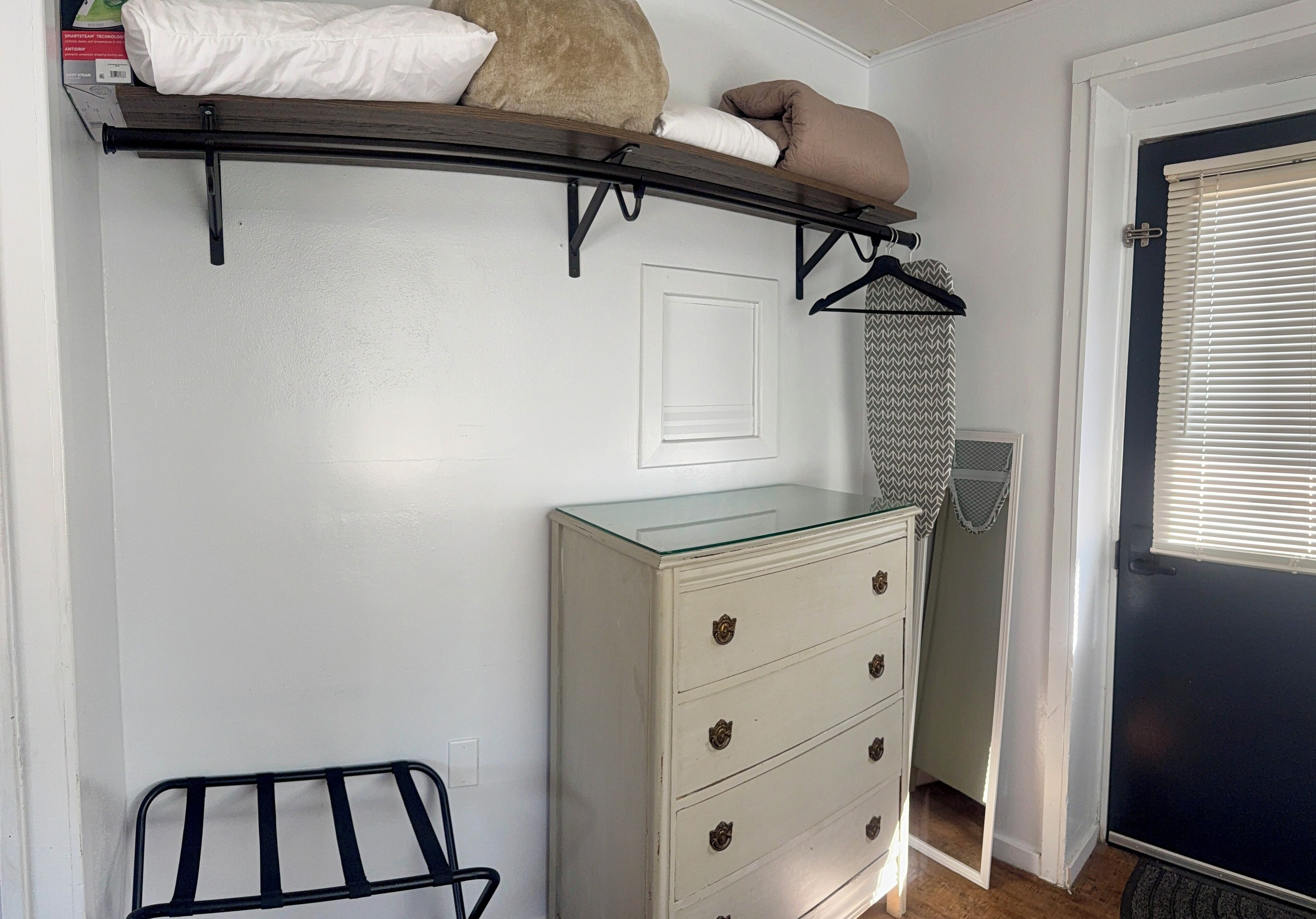 A room with a white dresser, a glass-top shelf with pillows, a black metal rack, and a door with a window.