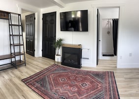 Una sala de estar con chimenea, televisor montado en la pared, piso de madera y alfombra estampada.