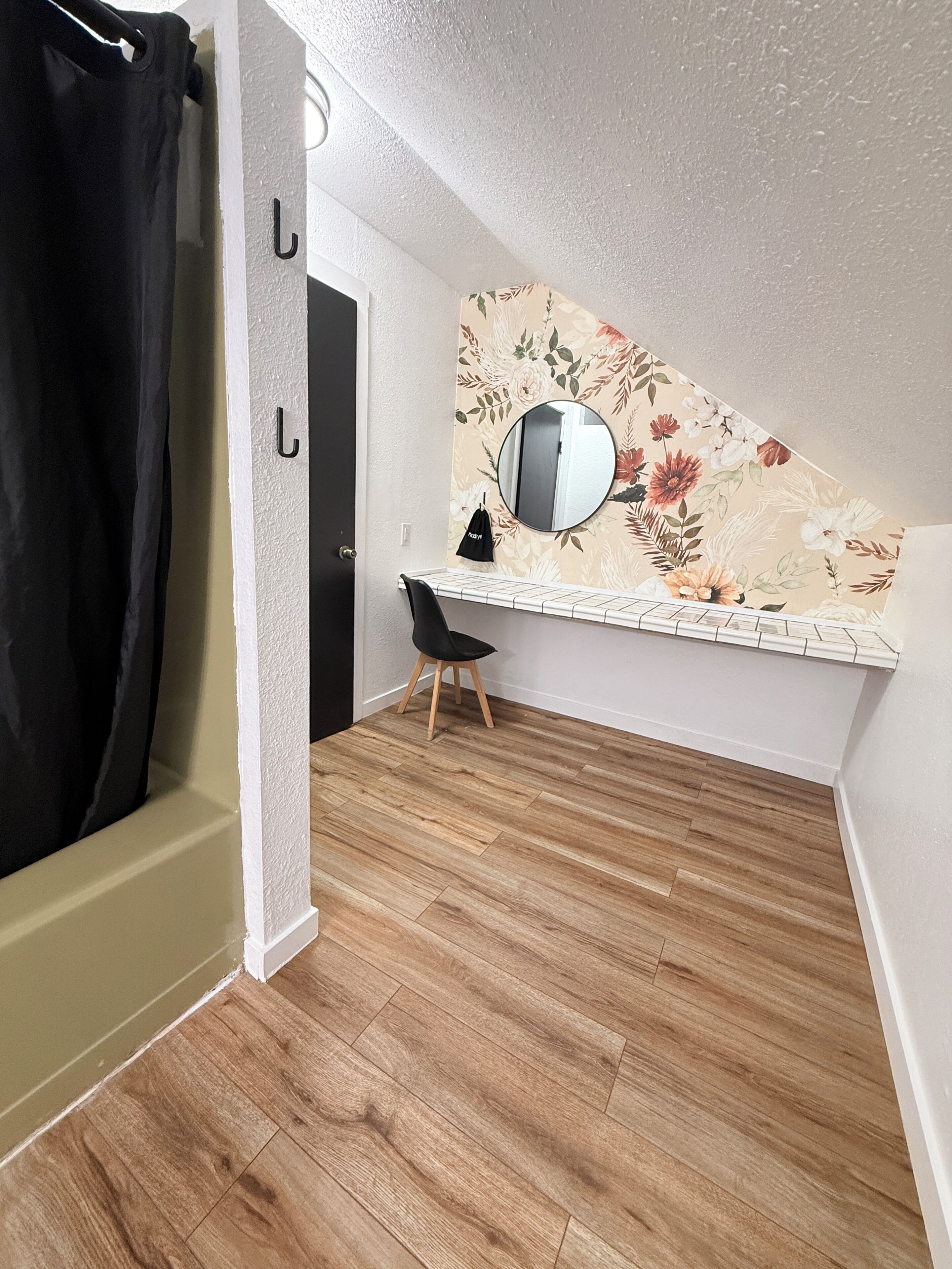 A room with a wooden floor, a black chair, a desk with a mirror, and a floral wallpaper.