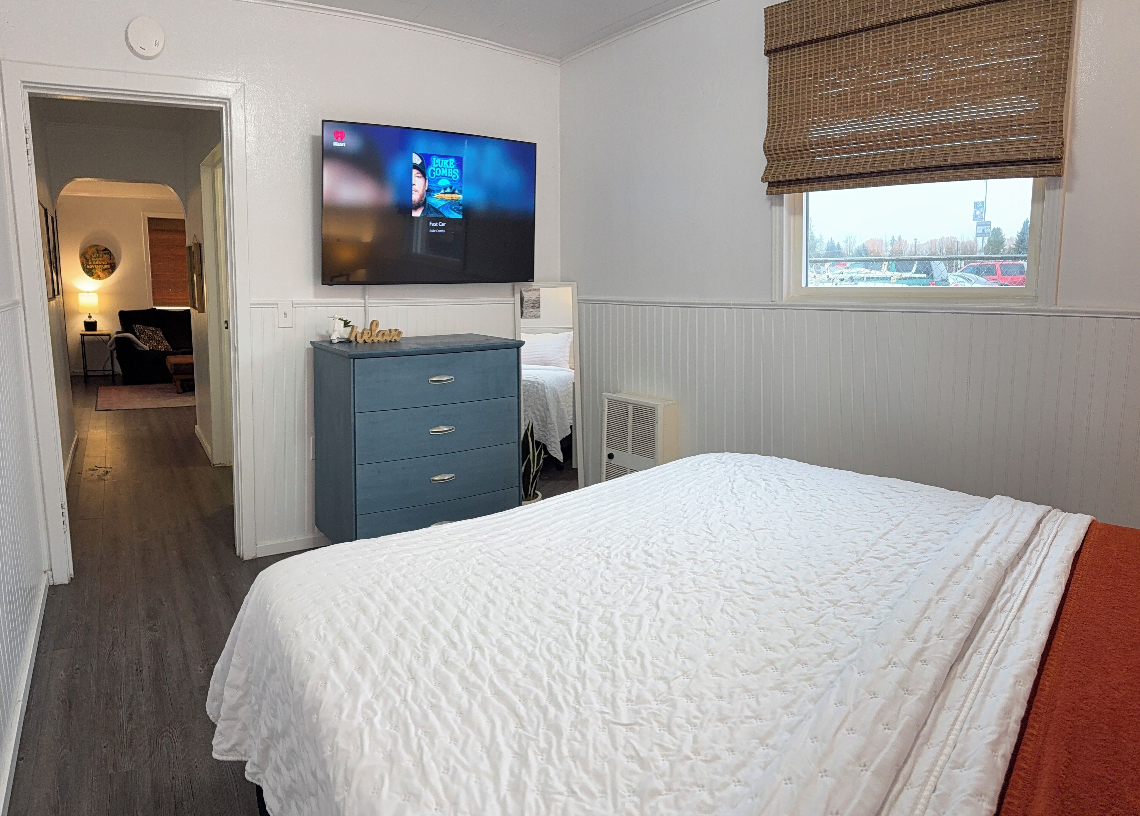 Een slaapkamer met een groot bed, een aan de muur bevestigde televisie, een dressoir en een raam met jaloezieën.