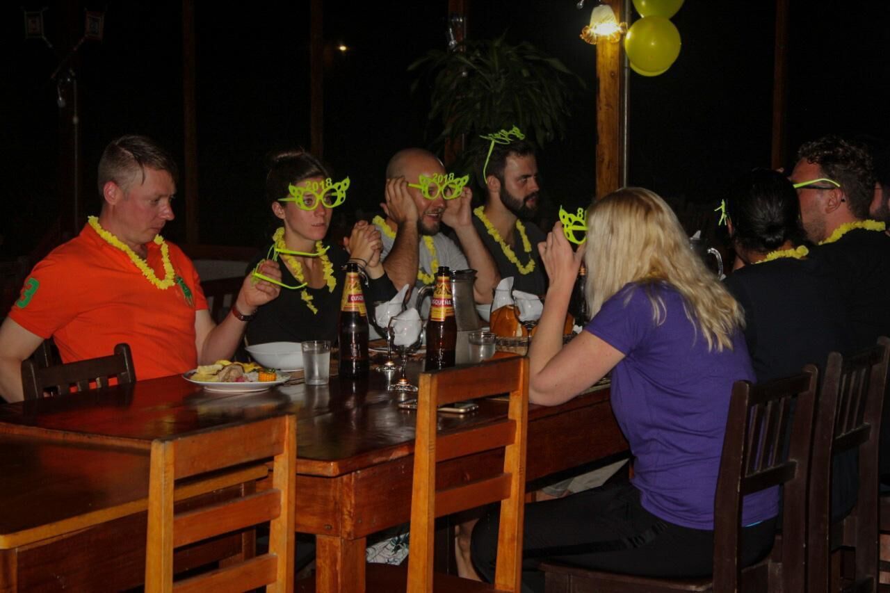 Een groep mensen zit rond een houten tafel met eten en drinken, versierd met feestelijke bloemkransen.
