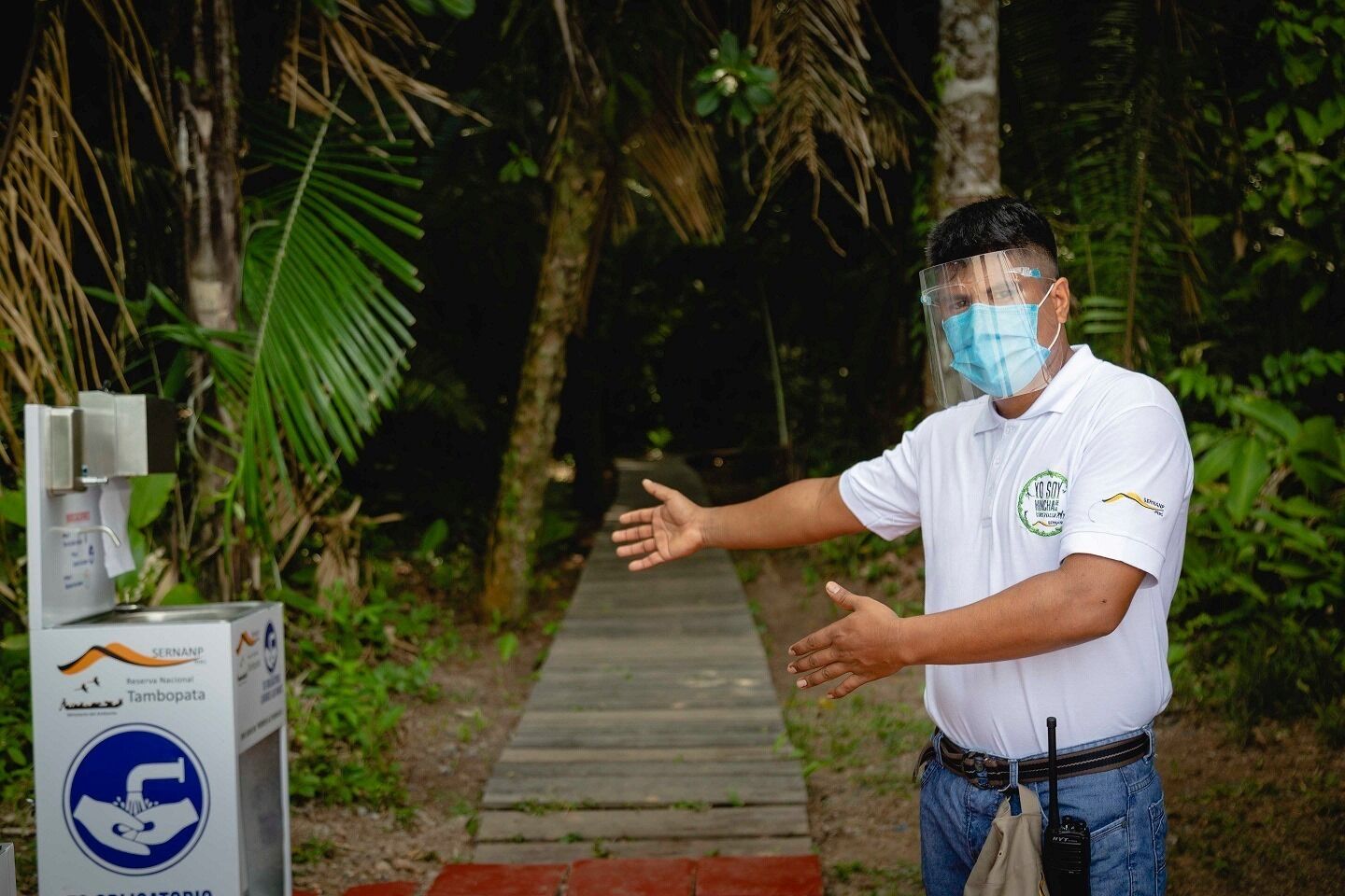 Un hombre con camisa blanca y un logo, gesticulando con las manos, de pie junto a un dispensador de agua con un letrero que dice "SERNANP Tambopata".