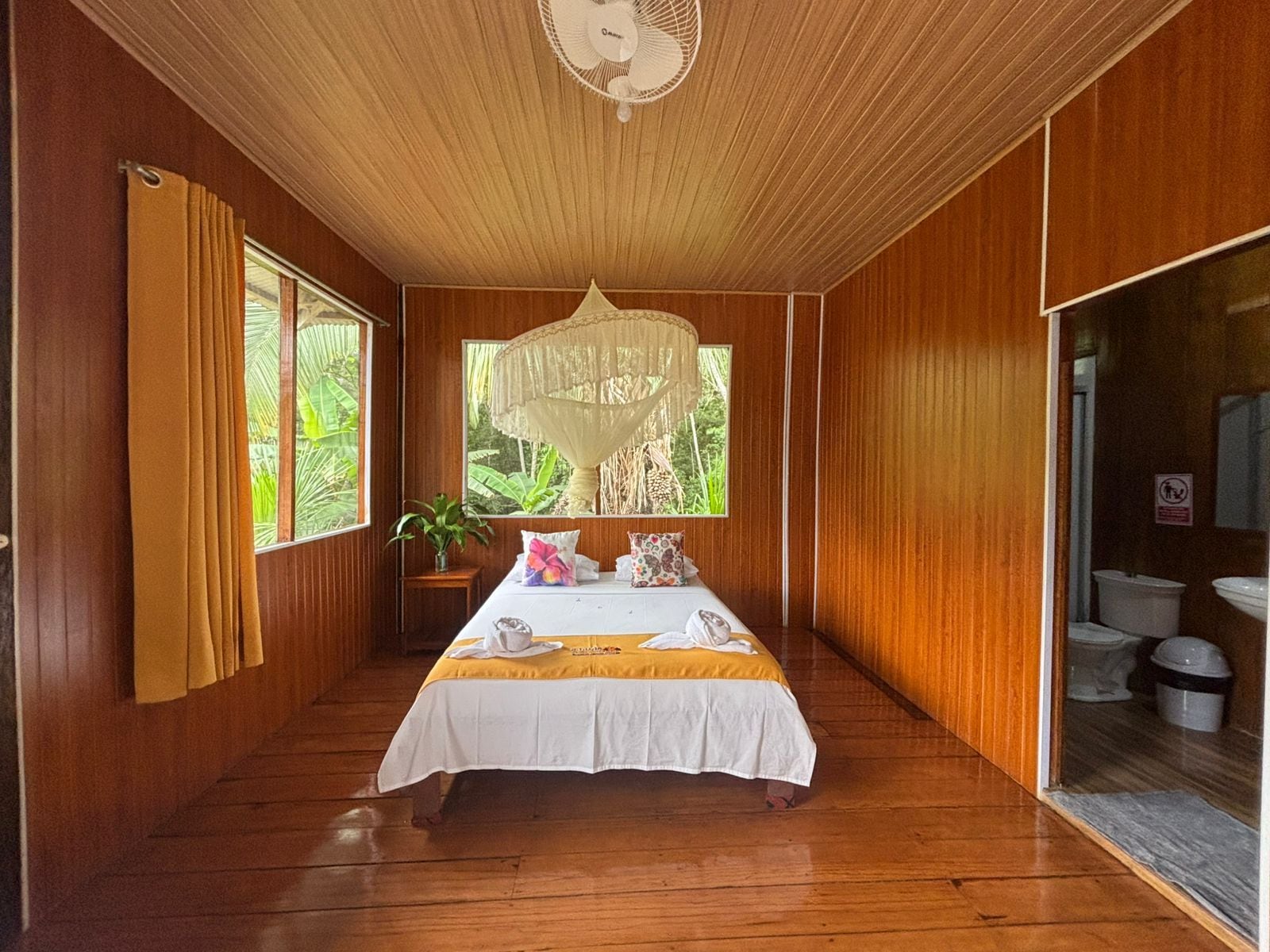 Une chambre avec un lit, un ventilateur de plafond, une fenêtre donnant sur un paysage verdoyant et une porte menant à la salle de bain.
