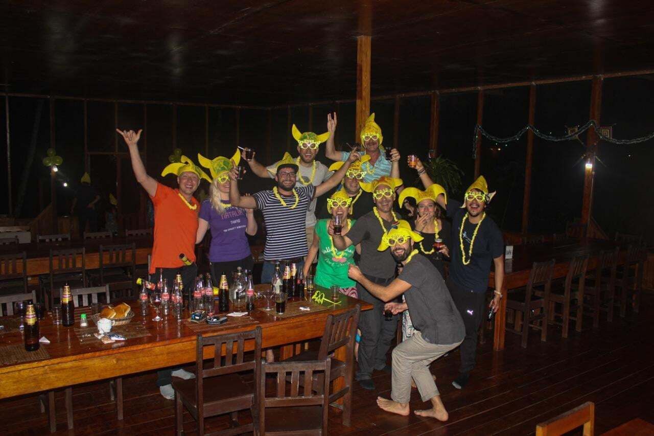 Um grupo de pessoas em uma mesa de madeira, com garrafas e copos, em um ambiente com piso e cadeiras de madeira.