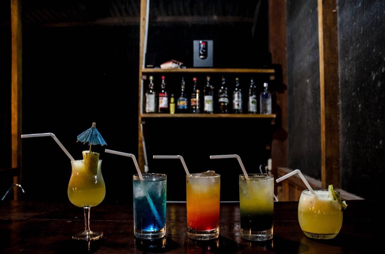 A bar with five colorful cocktails, each with a straw and a garnish. Behind the bar, there is a shelf with various bottles.
