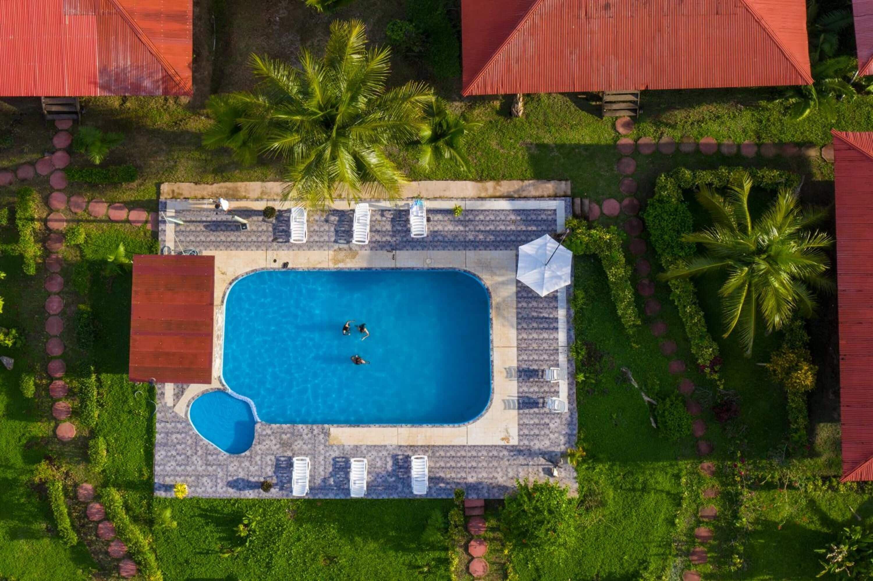 Vue aérienne d’un complexe hôtelier doté d’une piscine rectangulaire, entourée de chaises longues et de palmiers.