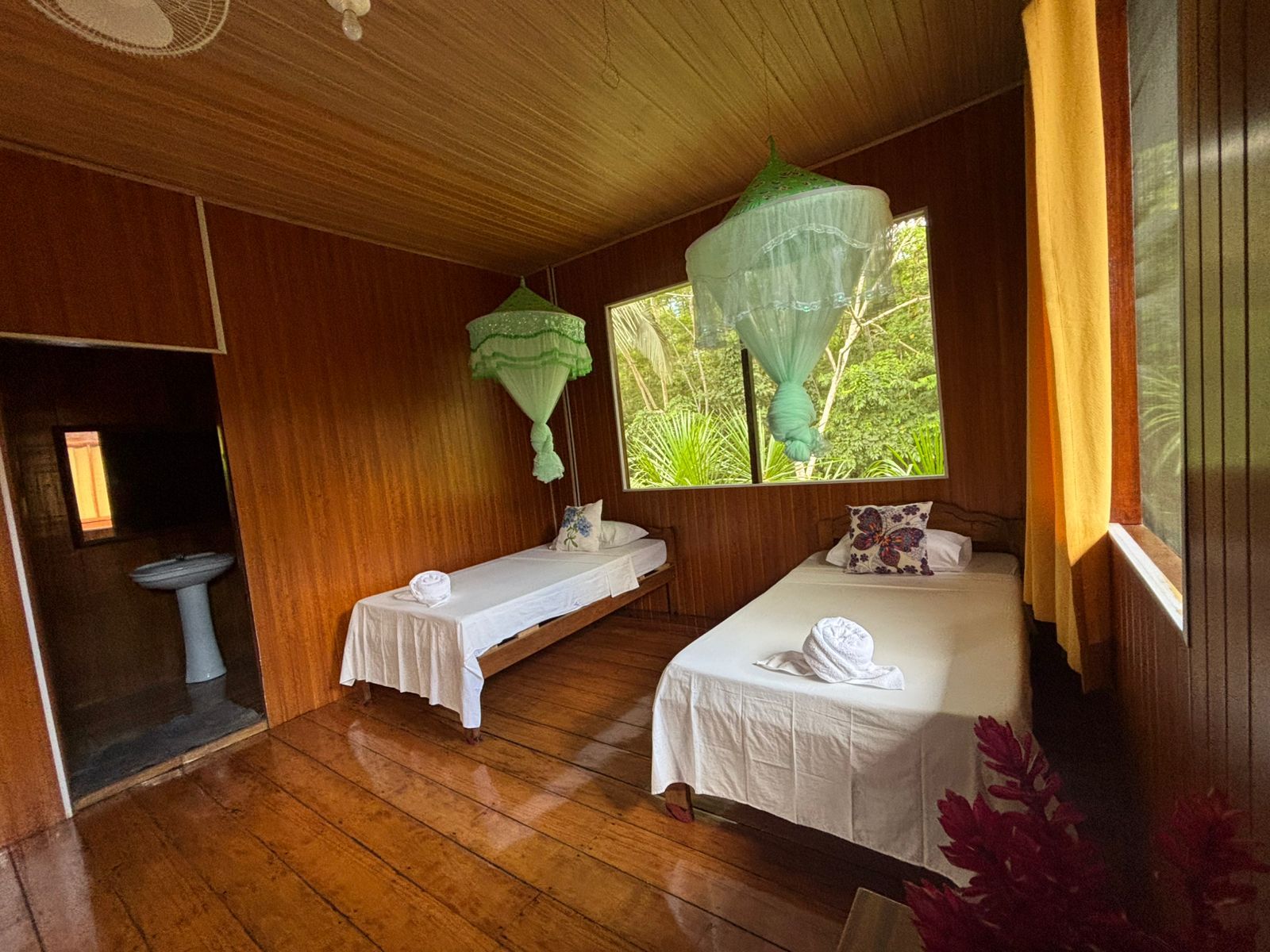 A room with two beds, a window with a view of greenery, a sink, and a ceiling fan.