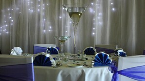 Una mesa dispuesta para un evento formal, con un centro de mesa alto de vidrio, lazos azules en las sillas y un mantel blanco.