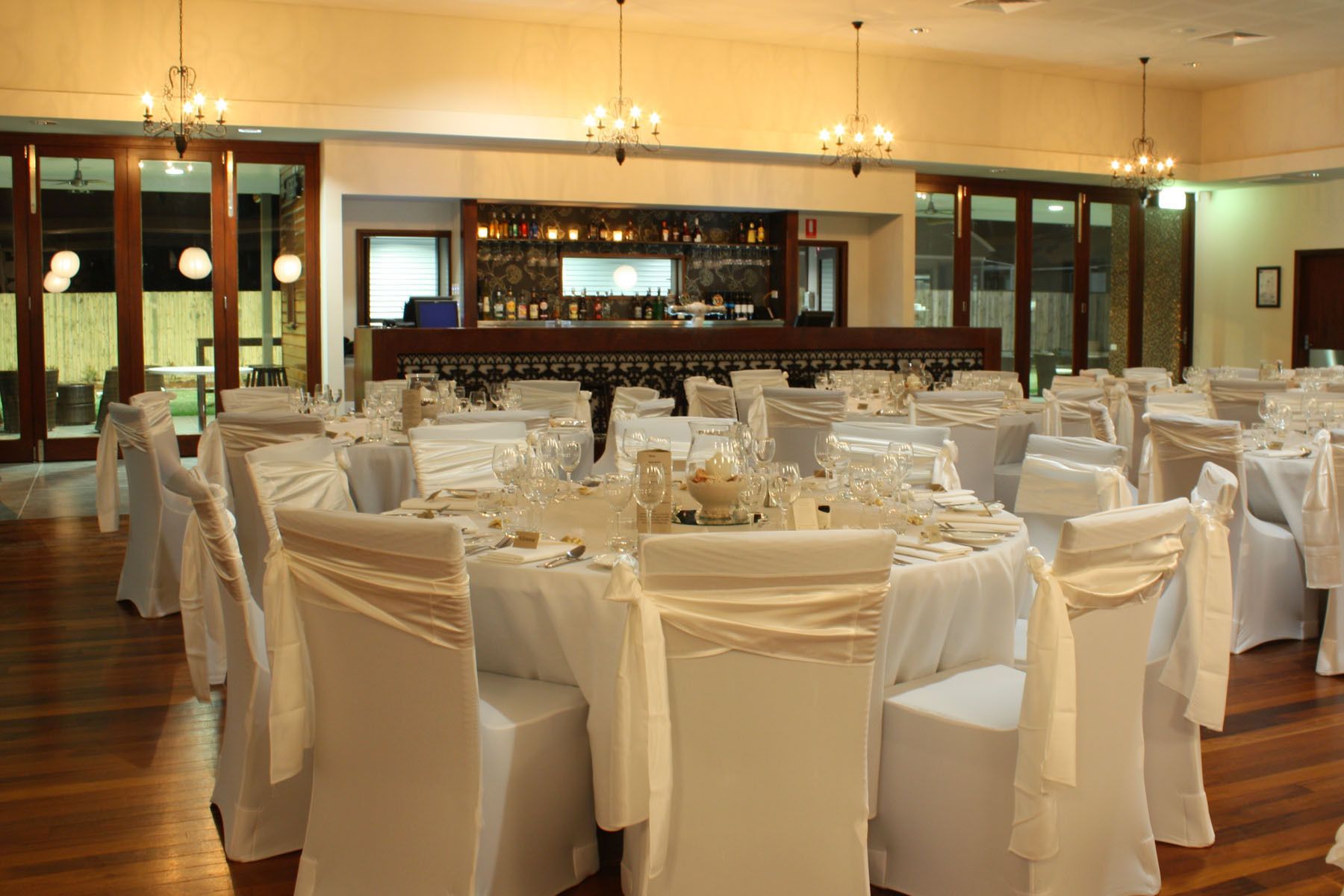 Un salón de banquetes con mesas redondas preparadas para un evento formal, una zona de bar con una variedad de botellas, y una araña colgando del techo.