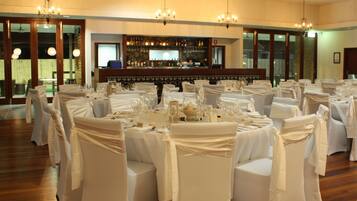 Un salón de banquetes con mesas redondas preparadas para un evento formal, una zona de bar con una variedad de botellas, y una araña colgando del techo.