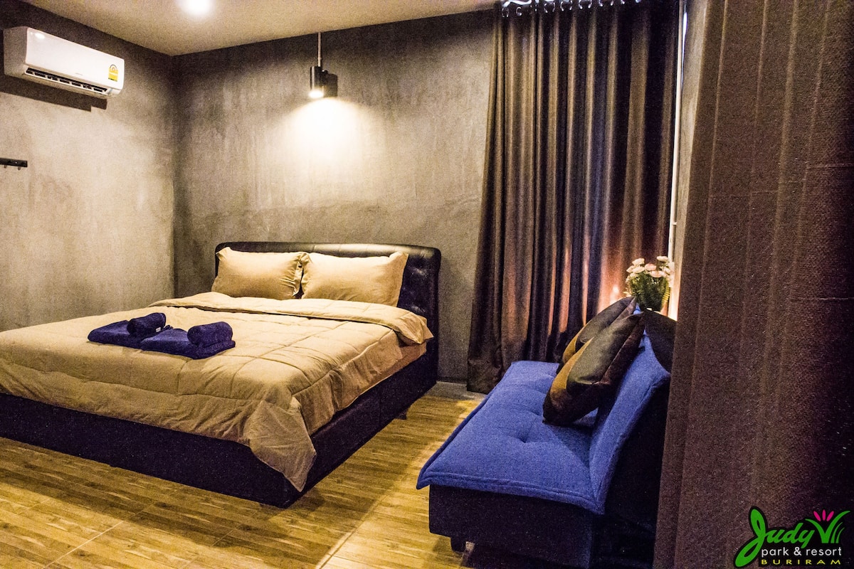 A bedroom with a bed, a bedside table, a hanging light, a window with curtains, and an air conditioning unit.