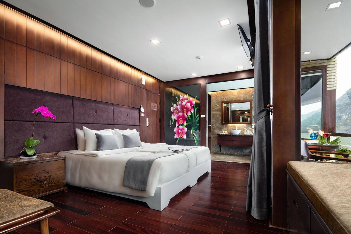 Een moderne slaapkamer met een groot bed, houten lambrisering, een nachtkastje, een bankje en een zichtbare badkamer door een open deur.