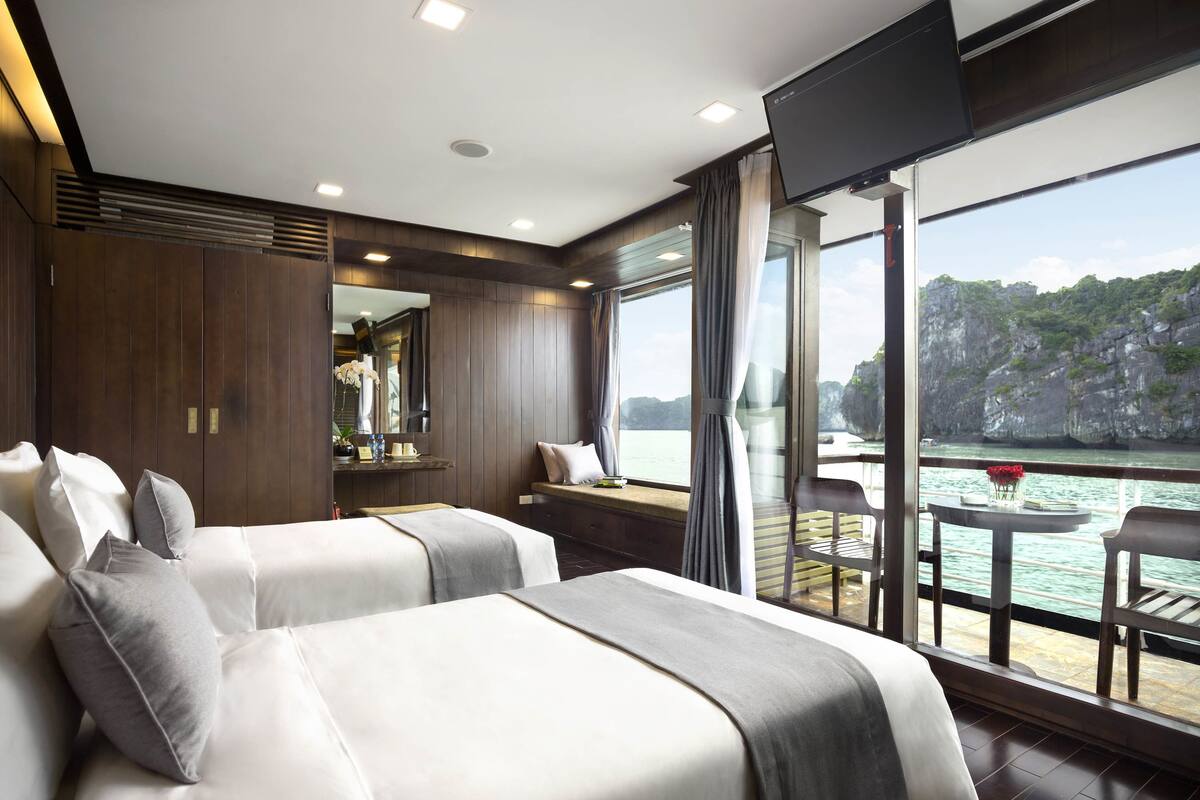 Een moderne hotelkamer met twee bedden, een groot raam met uitzicht op zee en een balkon met een tafel en stoelen.