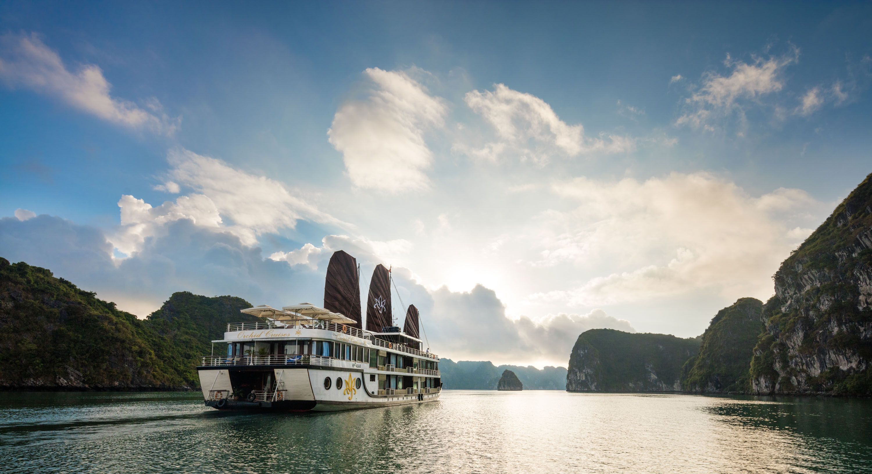 A river cruise ship with multiple decks and a logo on the side, surrounded by lush green hills and clear blue sky.
